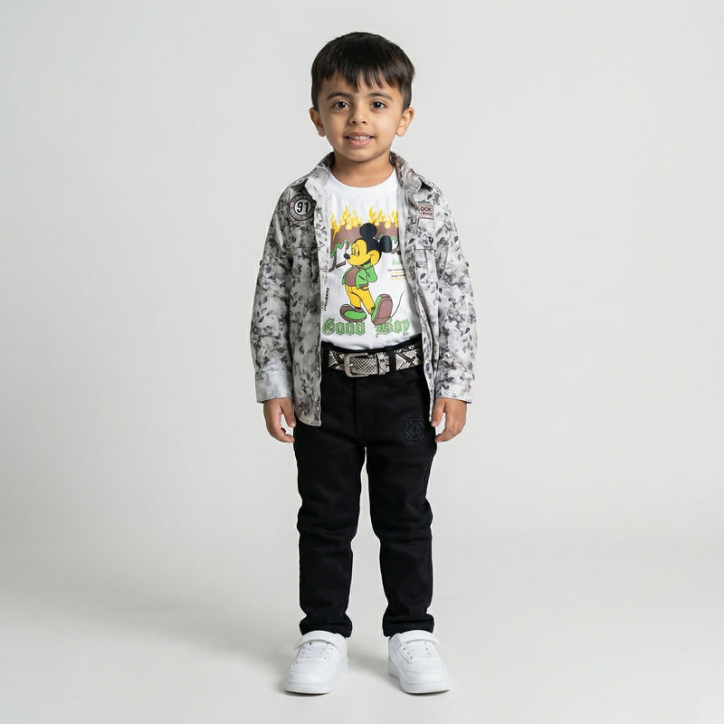 Young boy wearing a grey printed shirt over a white graphic t-shirt with black trousers, belt, and white sneakers against a light studio background