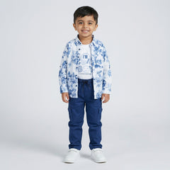 Young boy wearing a blue floral print shirt over a white graphic t-shirt with blue cargo pants and white sneakers against a light studio background