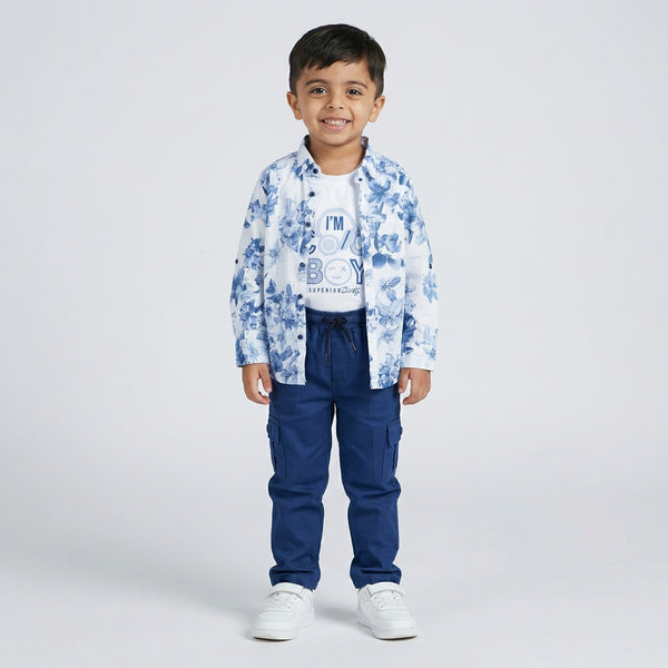 Young boy wearing a blue floral print shirt over a white graphic t-shirt with blue cargo pants and white sneakers against a light studio background