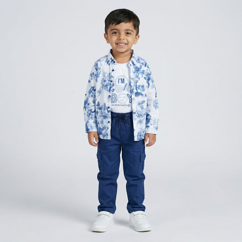Young boy wearing a blue floral print shirt over a white graphic t-shirt with blue cargo pants and white sneakers against a light studio background