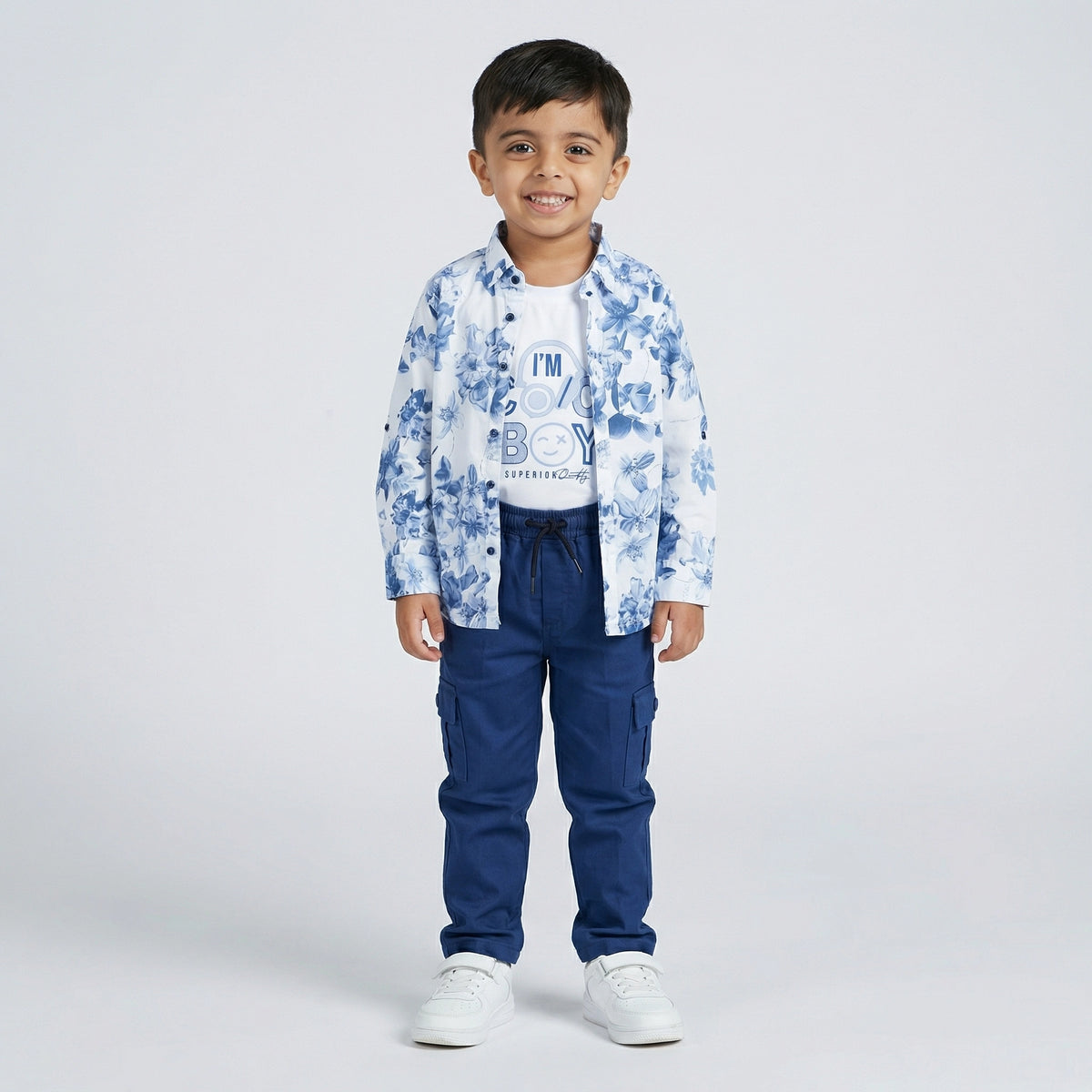 Young boy wearing a blue floral print shirt over a white graphic t-shirt with blue cargo pants and white sneakers against a light studio background