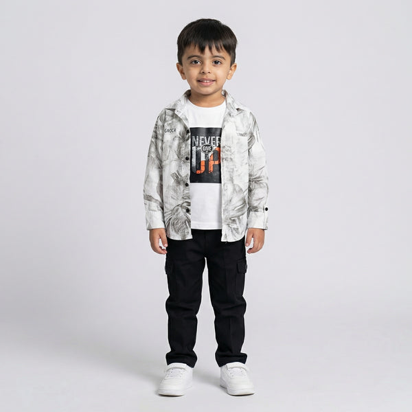 Young boy wearing a grey printed shirt over a white graphic t-shirt with black cargo pants and white sneakers against a light studio background