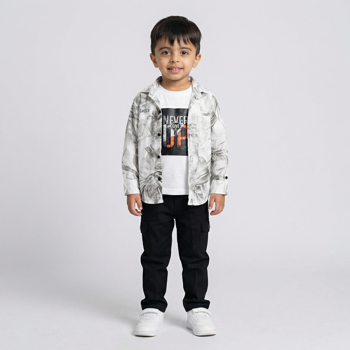 Young boy wearing a grey printed shirt over a white graphic t-shirt with black cargo pants and white sneakers against a light studio background