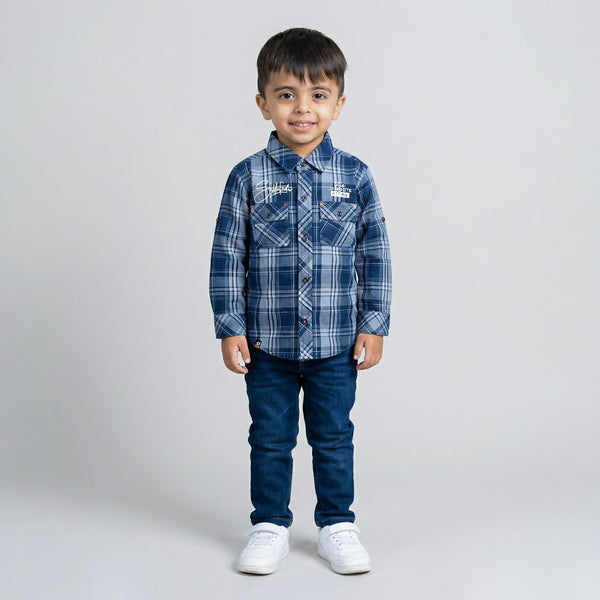 Young boy wearing a blue checked long-sleeve shirt with dark blue denim jeans and white sneakers against a light grey studio background