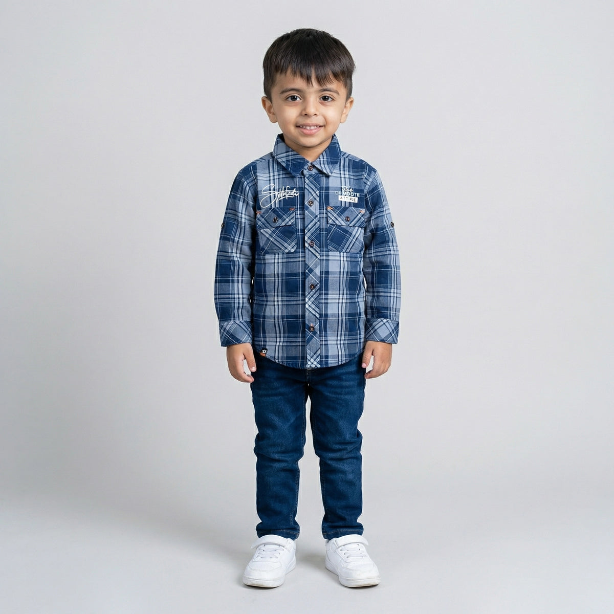 Young boy wearing a blue checked long-sleeve shirt with dark blue denim jeans and white sneakers against a light grey studio background