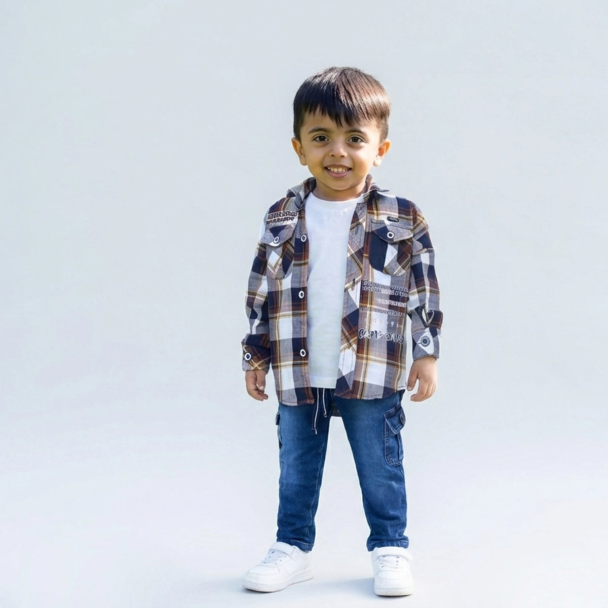 Young boy smiling in a brown checked casual shirt over a white T-shirt, blue cargo jeans, and white sneakers, standing against a plain light background.