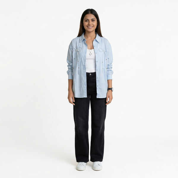 A woman wearing a light blue button-down shirt with bow embellishment details, layered over a white top, paired with black straight-leg jeans and white sneak