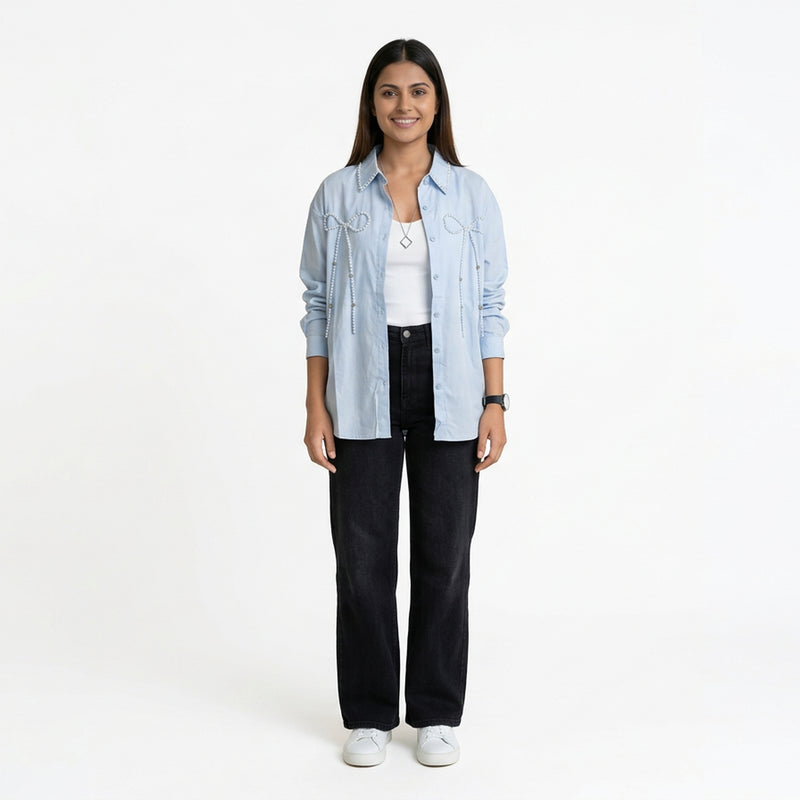 A woman wearing a light blue button-down shirt with bow embellishment details, layered over a white top, paired with black straight-leg jeans and white sneak