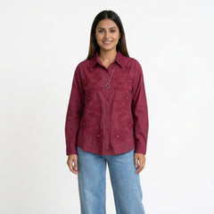 A woman wearing a maroon button-down shirt with intricate embroidered detailing and a textured collar, paired with light blue