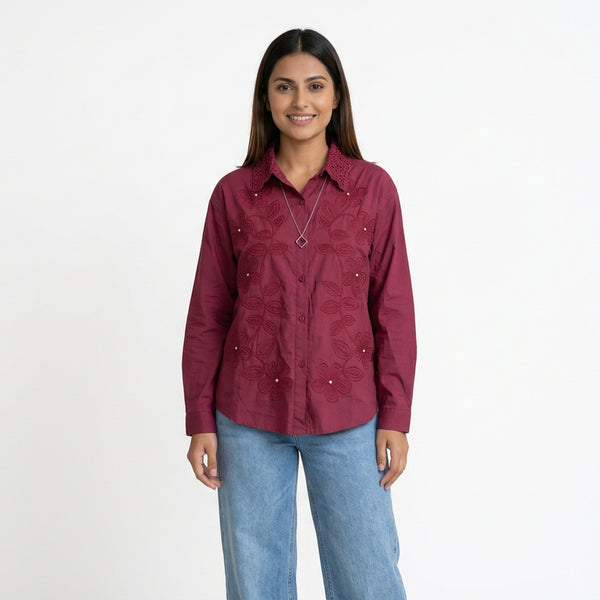 A woman wearing a maroon button-down shirt with intricate embroidered detailing and a textured collar, paired with light blue