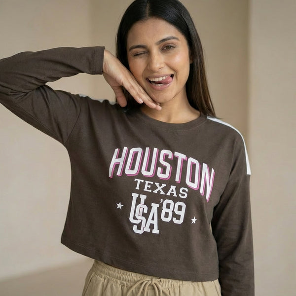 Woman wearing a brown cropped sweatshirt with “Houston Texas USA ’89” text, paired with beige drawstring pants, posing playfully with a wink and hand under chin.

