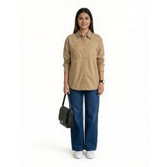 A woman wearing a beige button-down shirt with floral embroidery and studded collar detailing, paired with blue straight-leg jeans and white sneakers, holding a structured