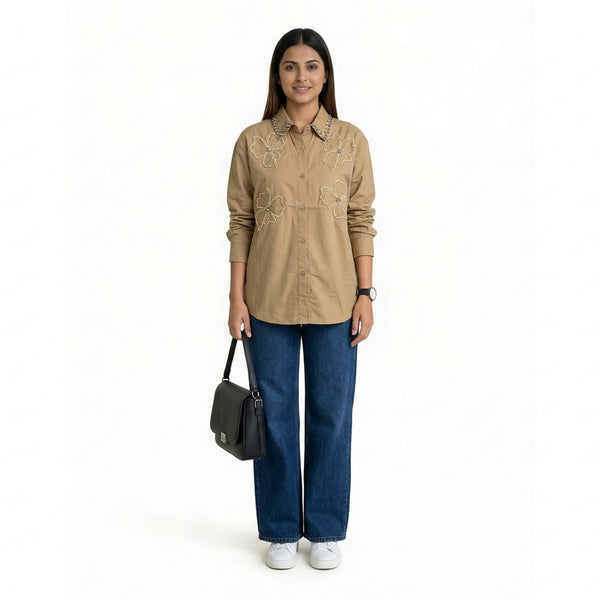 A woman wearing a beige button-down shirt with floral embroidery and studded collar detailing, paired with blue straight-leg jeans and white sneakers, holding a structured