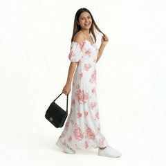 A smiling woman wearing a white maxi dress with pink floral prints and off-shoulder sleeves, paired with white sneakers. She is holding a black handbag and posing casually while looking at the camera against a plain white background.