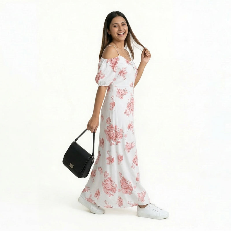 A smiling woman wearing a white maxi dress with pink floral prints and off-shoulder sleeves, paired with white sneakers. She is holding a black handbag and posing casually while looking at the camera against a plain white background.
