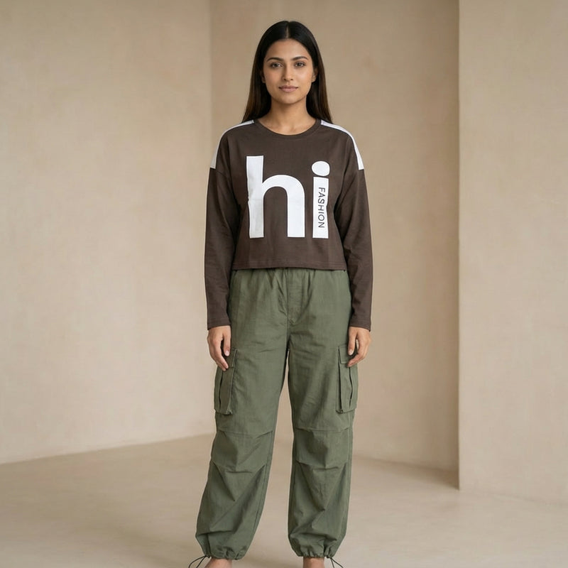 Woman wearing a brown cropped long-sleeve top with “hi fashion” graphic text, paired with olive green cargo pants, standing in a minimal indoor se

