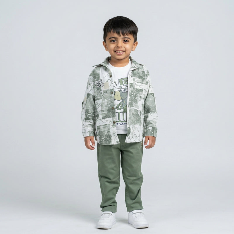 Young boy wearing a sage green printed shirt over a white graphic t-shirt with matching green pants and white sneakers against a light studio background
