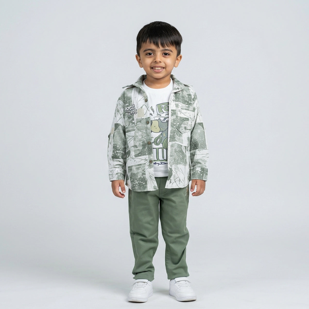 Young boy wearing a sage green printed shirt over a white graphic t-shirt with matching green pants and white sneakers against a light studio background