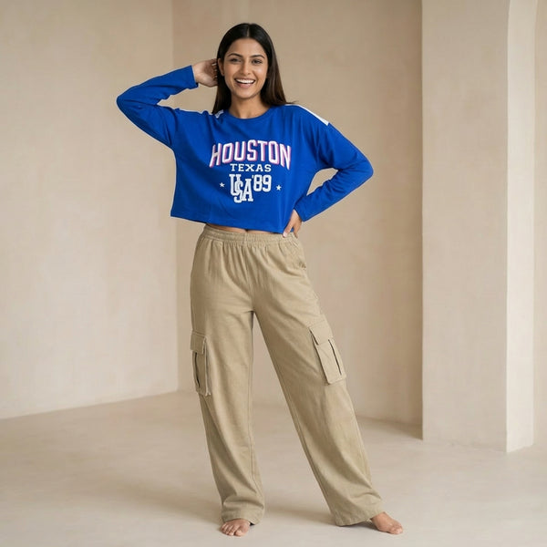 Woman wearing a blue cropped sweatshirt with “Houston Texas USA ’89” text, paired with beige cargo pants, posing casually with a hand on her head in a minimal indoor setting.

