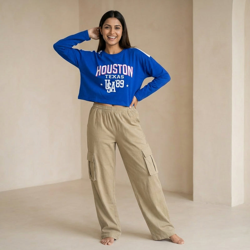 Woman wearing a blue cropped sweatshirt with “Houston Texas USA ’89” text, paired with beige cargo pants, posing casually with a hand on her head in a minimal indoor setting.

