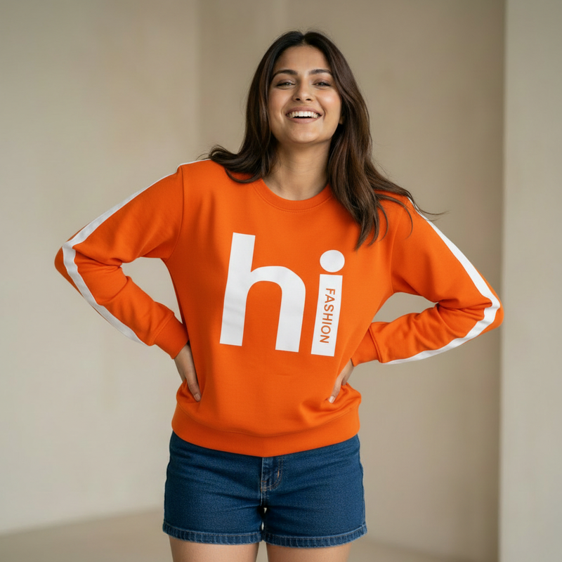 Woman wearing an orange sweatshirt with “hi fashion” graphic text and white sleeve stripes, paired with denim shorts, smiling in a minimal indoor setting.

