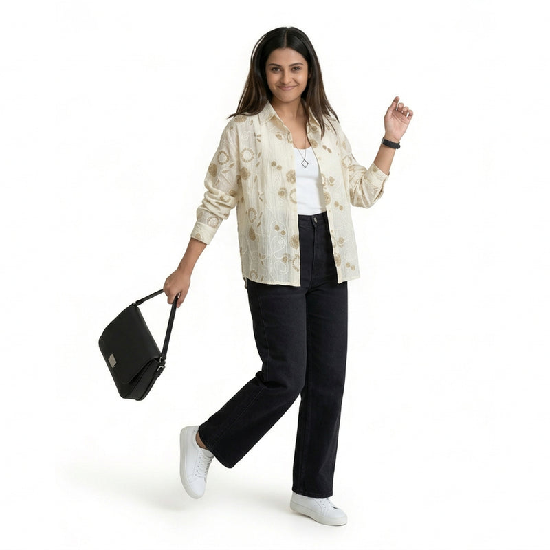 A woman wearing a cream printed button-down shirt over a white top, paired with dark wide-leg jeans and white sneakers, holding a black handbag and posing against a plain background.