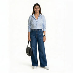 A smiling woman wearing a light blue button-down shirt with floral embroidery and studded collar detailing, paired with blue wide-leg jeans and white sneakers, holding a structured black handbag.