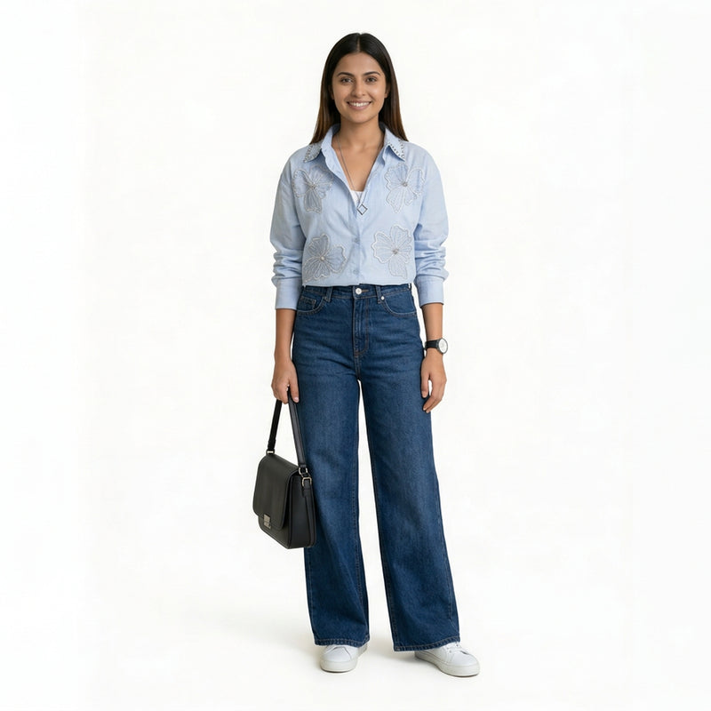 A smiling woman wearing a light blue button-down shirt with floral embroidery and studded collar detailing, paired with blue wide-leg jeans and white sneakers, holding a structured black handbag.