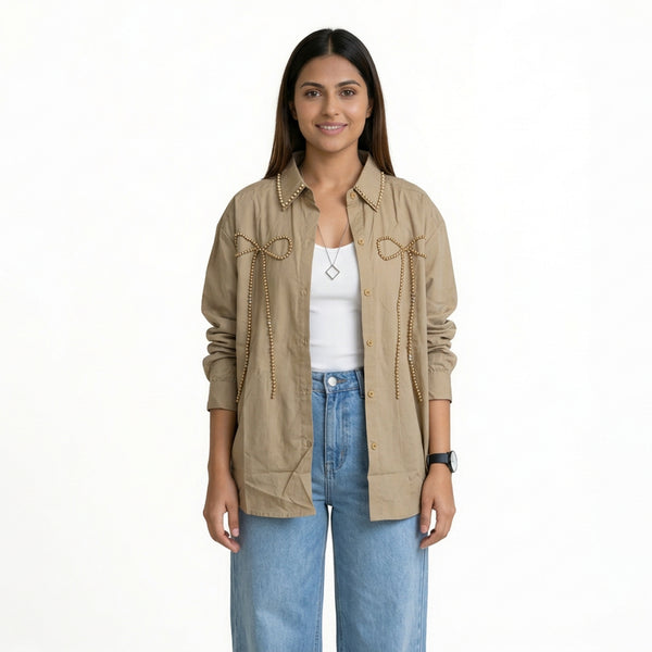 A woman wearing a beige button-down shirt with bow embellishments and studded detailing, layered over a white top, paired with light blue