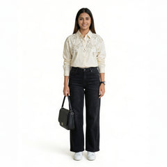 A woman wearing a cream embroidered button-down shirt paired with black straight-leg jeans, white sneakers, and carrying a black shoulder bag.

