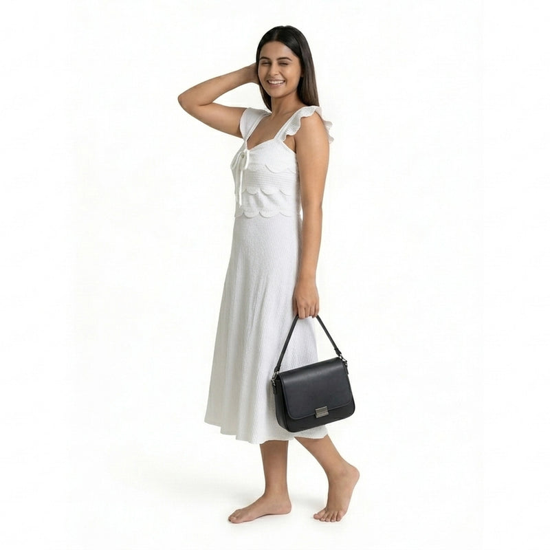 A smiling woman wearing a sleeveless off-white midi dress with ruffled straps and a textured finish, posing barefoot while holding a structured black handbag.