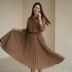 Woman wearing a brown shirt-style dress with a cinched waist and pleated skirt, holding the hem while standing in a minimal indoor settin

