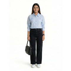 A woman wearing a light blue button-down shirt with floral embroidery and a textured collar, paired with black straight-leg jeans and white sneakers, holding a structured black handbag.