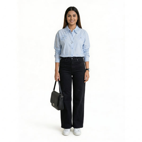 A woman wearing a light blue button-down shirt with floral embroidery and a textured collar, paired with black straight-leg jeans and white sneakers, holding a structured black handbag.