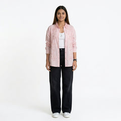 A woman wearing a light pink button-down shirt with delicate embroidered detailing, layered over a white top, paired with black straight-leg jeans and white sneakers.