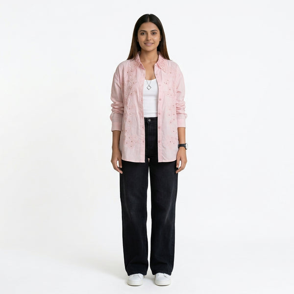A woman wearing a light pink button-down shirt with delicate embroidered detailing, layered over a white top, paired with black straight-leg jeans and white sneakers.