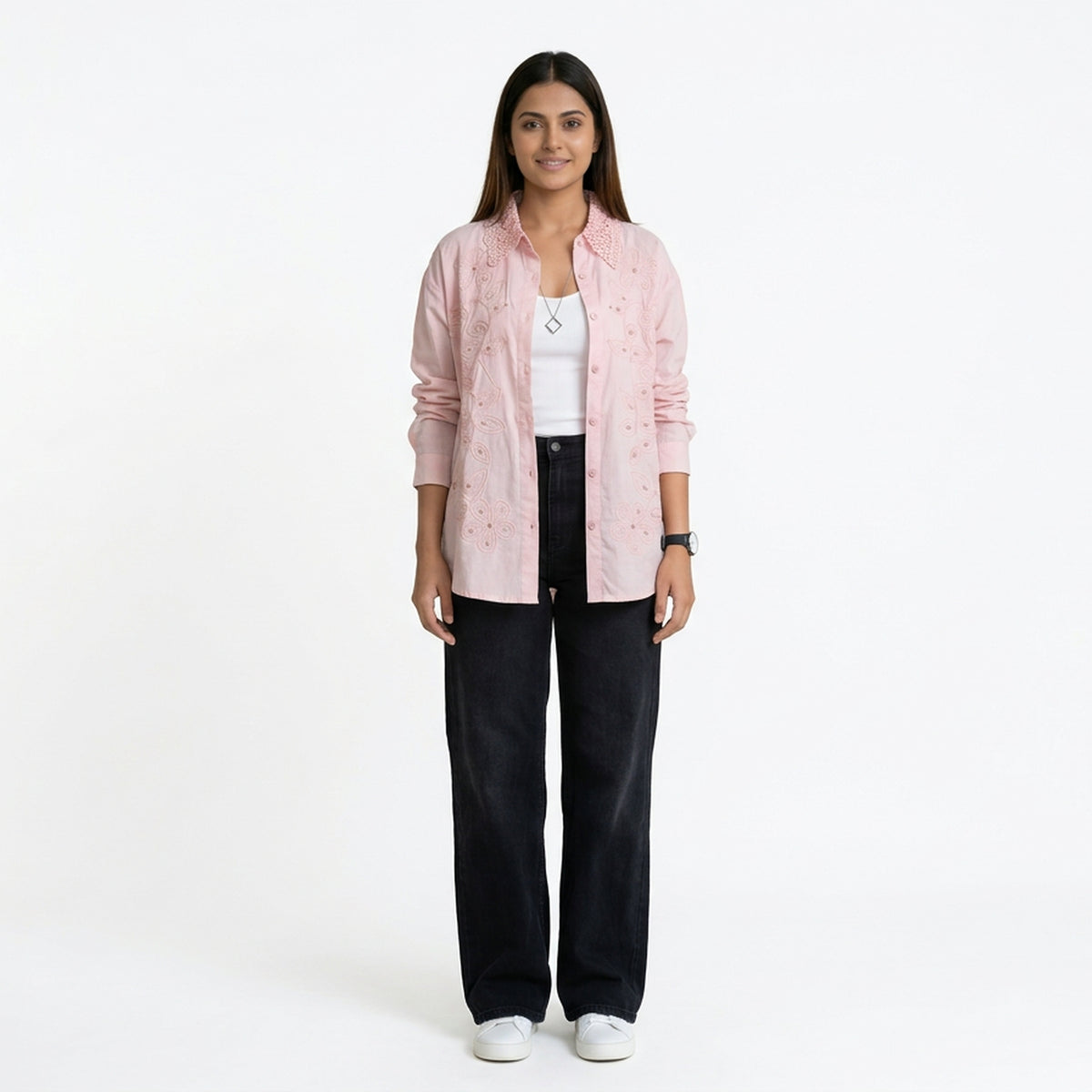A woman wearing a light pink button-down shirt with delicate embroidered detailing, layered over a white top, paired with black straight-leg jeans and white sneakers.