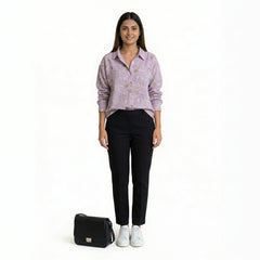 A woman wearing a lavender printed button-down shirt paired with slim black pants and white sneakers, with a black handbag placed beside her, against a plain backgro

