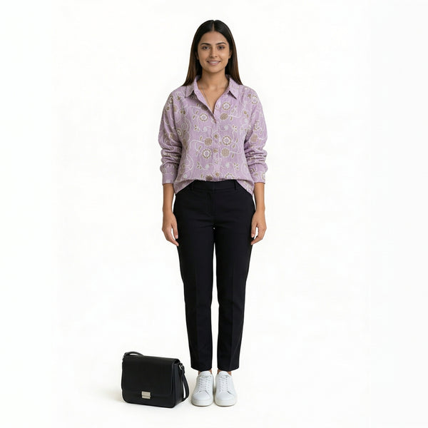 A woman wearing a lavender printed button-down shirt paired with slim black pants and white sneakers, with a black handbag placed beside her, against a plain backgro


