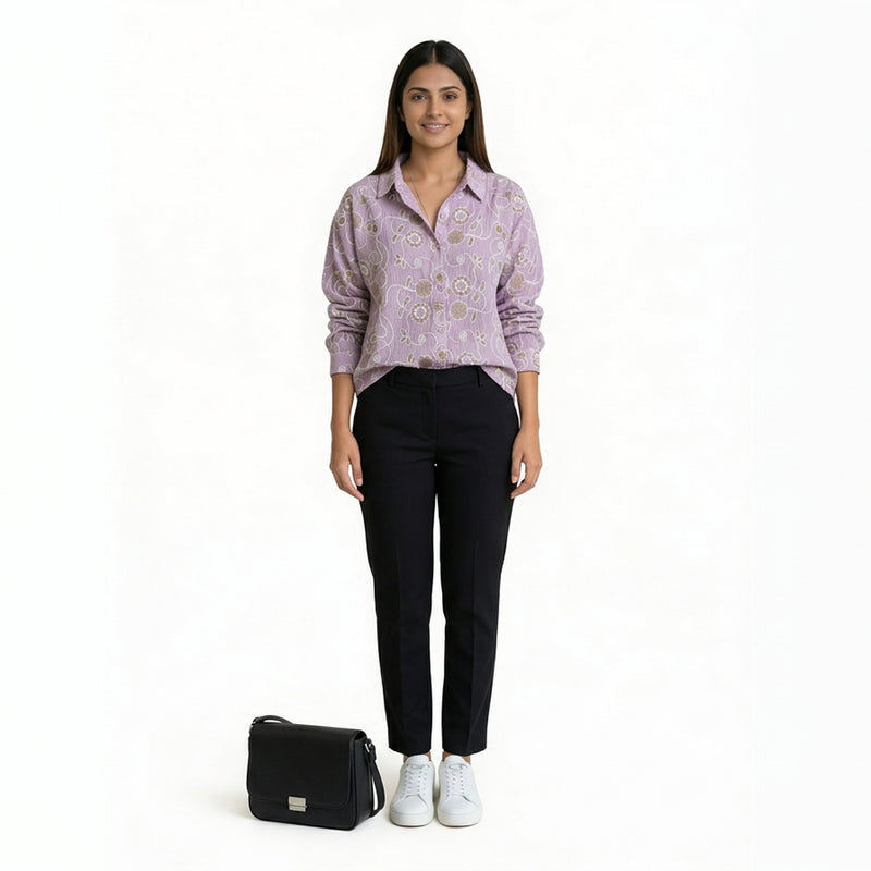 A woman wearing a lavender printed button-down shirt paired with slim black pants and white sneakers, with a black handbag placed beside her, against a plain backgro

