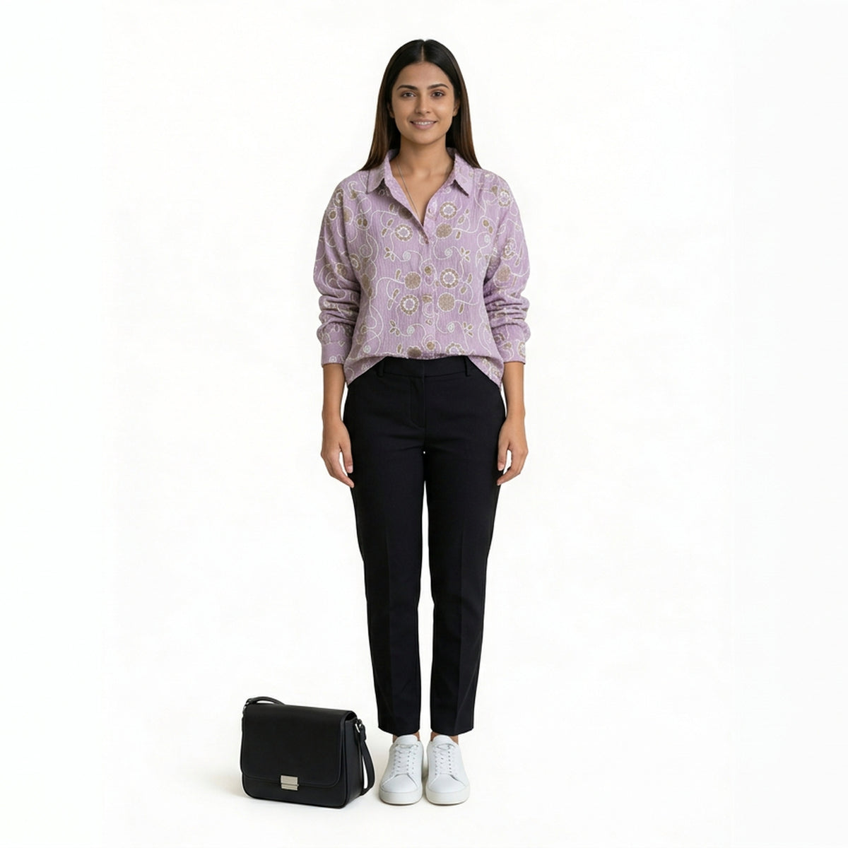 A woman wearing a lavender printed button-down shirt paired with slim black pants and white sneakers, with a black handbag placed beside her, against a plain backgro

