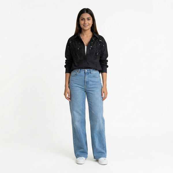 A woman wearing a black embellished button-down shirt paired with light blue straight-leg jeans and white sneakers.





