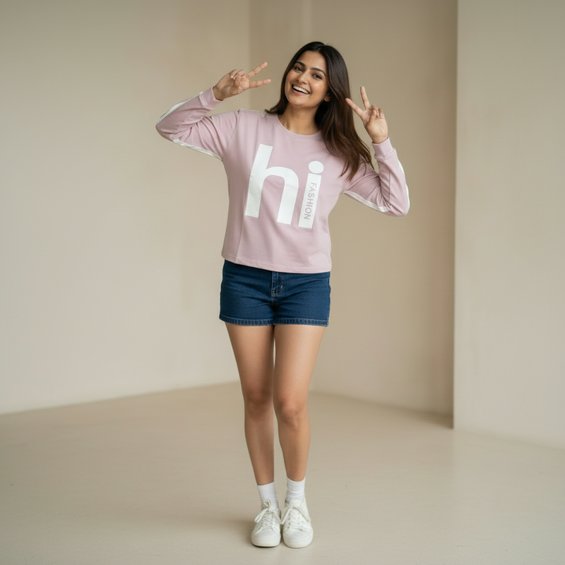 Woman wearing a light pink sweatshirt with “hi fashion” graphic print, paired with denim shorts and white sneakers, smiling and posing with peace signs in a minimal indoor setting.

