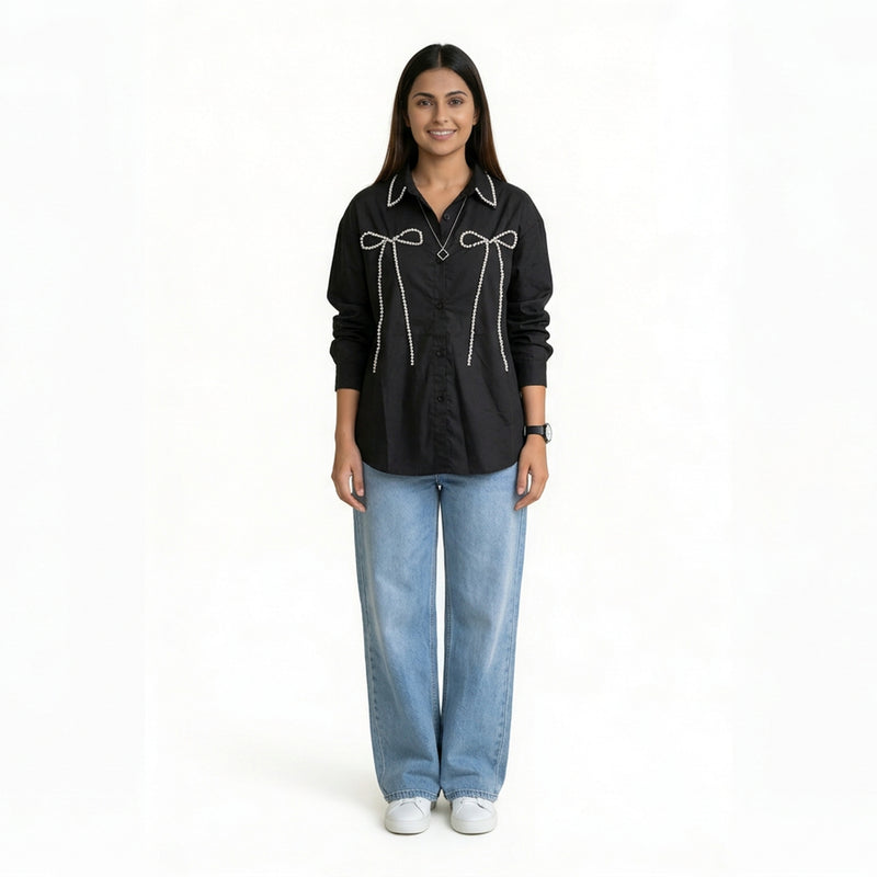A woman wearing a black button-down shirt with bow embellishment details on the front, paired with light blue wide-leg jeans and white sneakers.