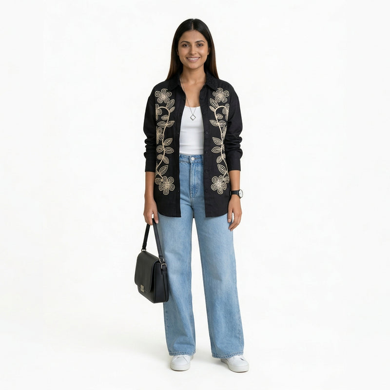 A smiling woman wearing a black button-down shirt with floral embroidery, layered over a white top, paired with light blue wide-leg jeans and white sneakers, holding a structured bla