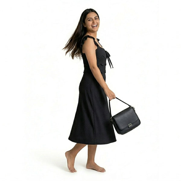A smiling woman wearing a sleeveless black midi dress with ruffled straps and a textured finish, walking barefoot while holding a structured black handbag.