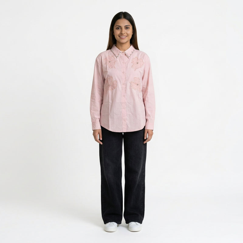 A woman wearing a light pink button-down shirt with floral embroidery and a studded collar, paired with dark blue wide-leg jeans and white sneakers.