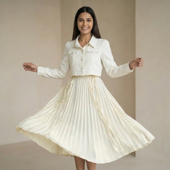 Woman wearing an off-white cropped shirt paired with a matching pleated midi skirt, twirling in a minimal indoor setting.

