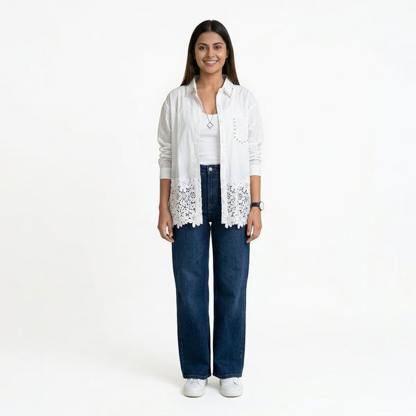 A smiling woman wearing a white button-down shirt with lace detailing at the hem and stud accents, layered over a white top, paired with blue wide-leg jeans and white sneakers.