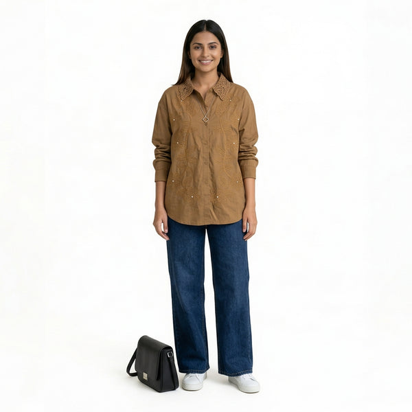 A woman wearing a brown button-down shirt with intricate embroidered detailing and a textured collar, paired with blue wide-leg jeans and white sneakers, with a black handbag placed beside h